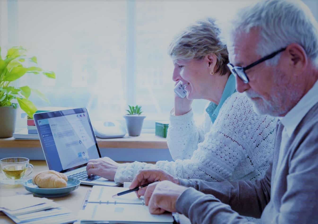 Senior Couple at Kitchen Table Completing Retirement Planning Checklist with laptop and pads of paper
